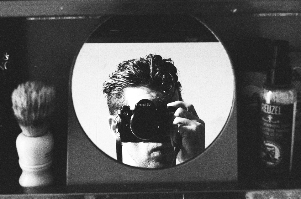 A black-and-white photo of a person taking a self-portrait in a round mirror, holding a camera in front of their face. Toiletries like a brush and a bottle are visible beside the mirror.